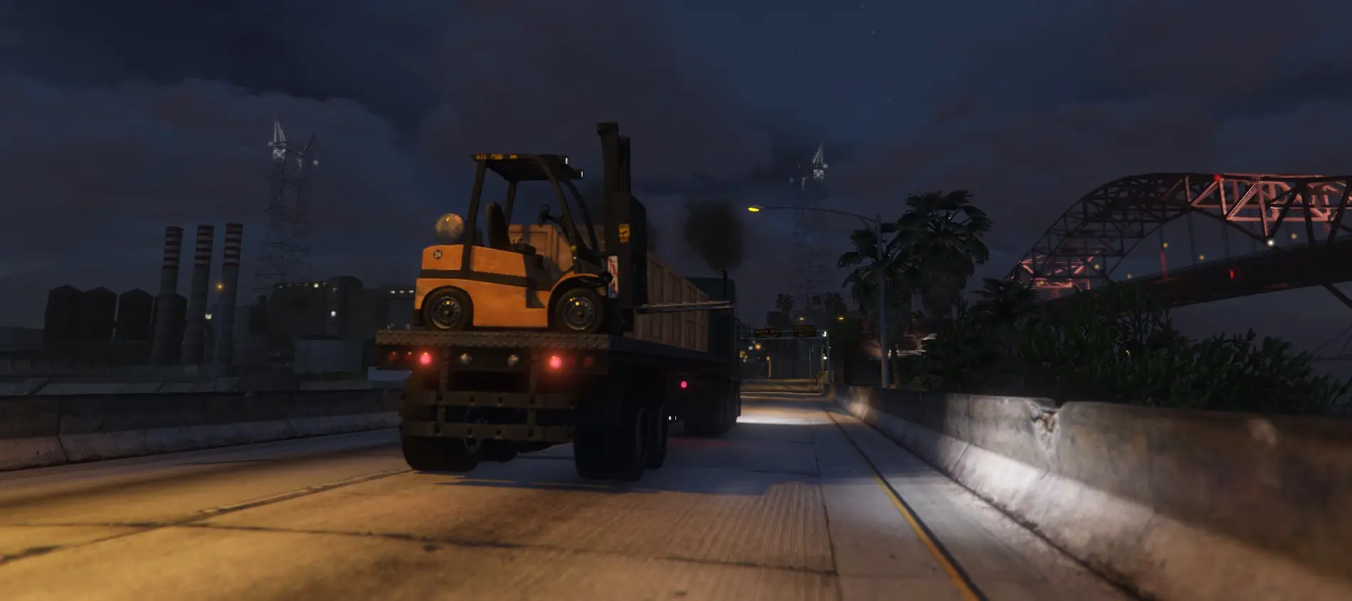 A flatbed truck carrying a forklift along a lit highway at night